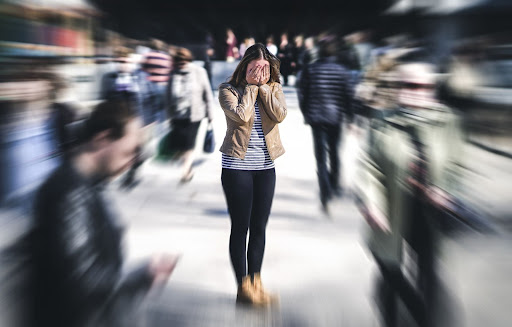 A woman standing with her hands over her eyes in the middle of a street. The surrounding people are blurred out.